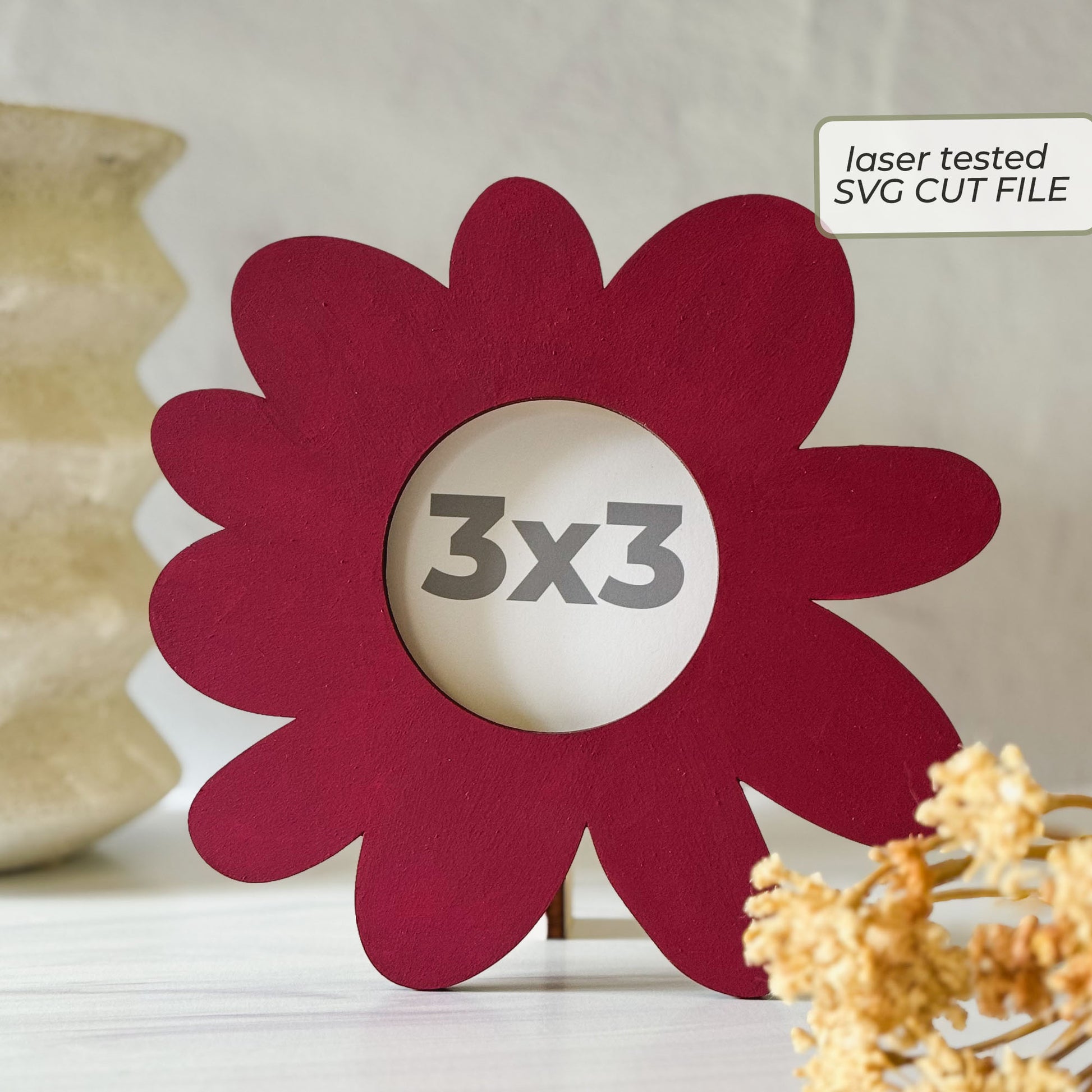 A flower-shaped red laser-cut picture frame with a 3x3 inch center cutout, displayed on a tabletop with decorative dried flowers and a vase in the background. A label in the top right reads 'laser tested SVG CUT FILE'.