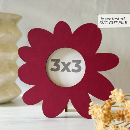 A flower-shaped red laser-cut picture frame with a 3x3 inch center cutout, displayed on a tabletop with decorative dried flowers and a vase in the background. A label in the top right reads 'laser tested SVG CUT FILE'.
