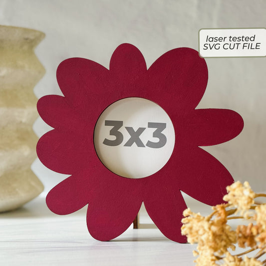 A flower-shaped red laser-cut picture frame with a 3x3 inch center cutout, displayed on a tabletop with decorative dried flowers and a vase in the background. A label in the top right reads 'laser tested SVG CUT FILE'.