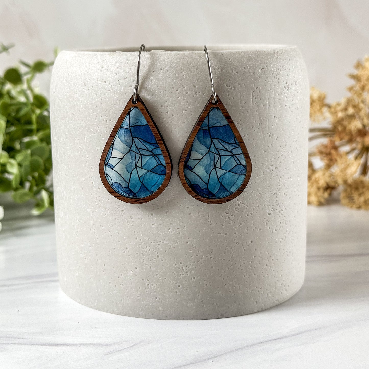 Teardrop-shaped blue and brown earrings on a white cylindrical holder with a light background.