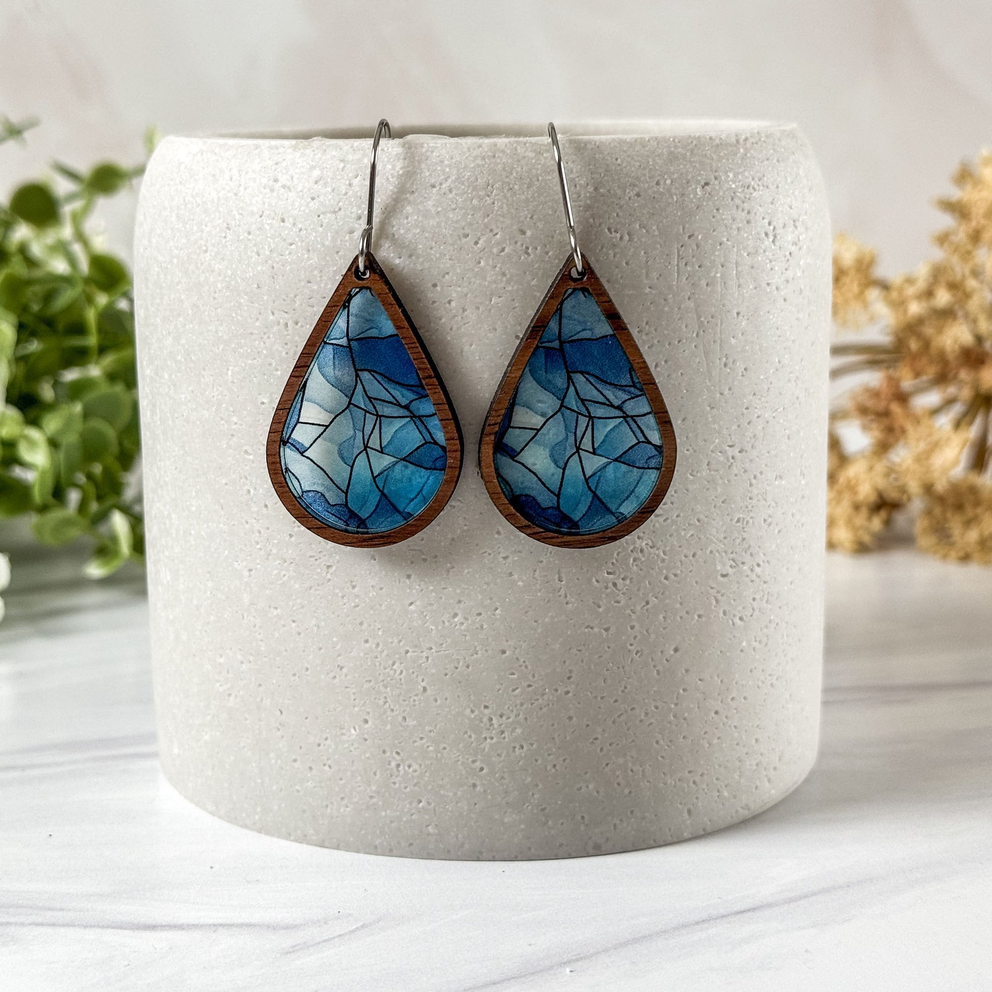 Teardrop-shaped blue and brown earrings on a white cylindrical holder with a light background.