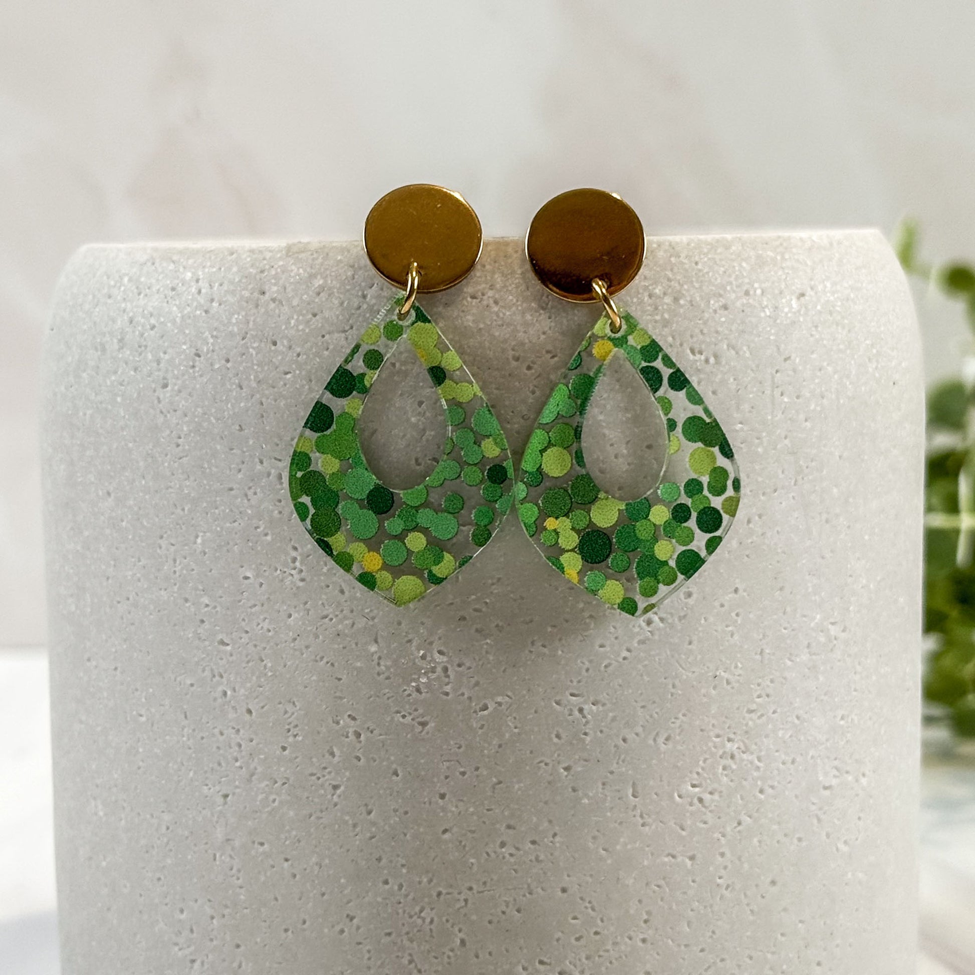 Green patterned earrings on a white cylindrical stand with a blurred green plant in the background.