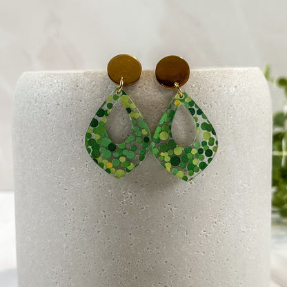 Green patterned earrings on a white cylindrical stand with a blurred green plant in the background.