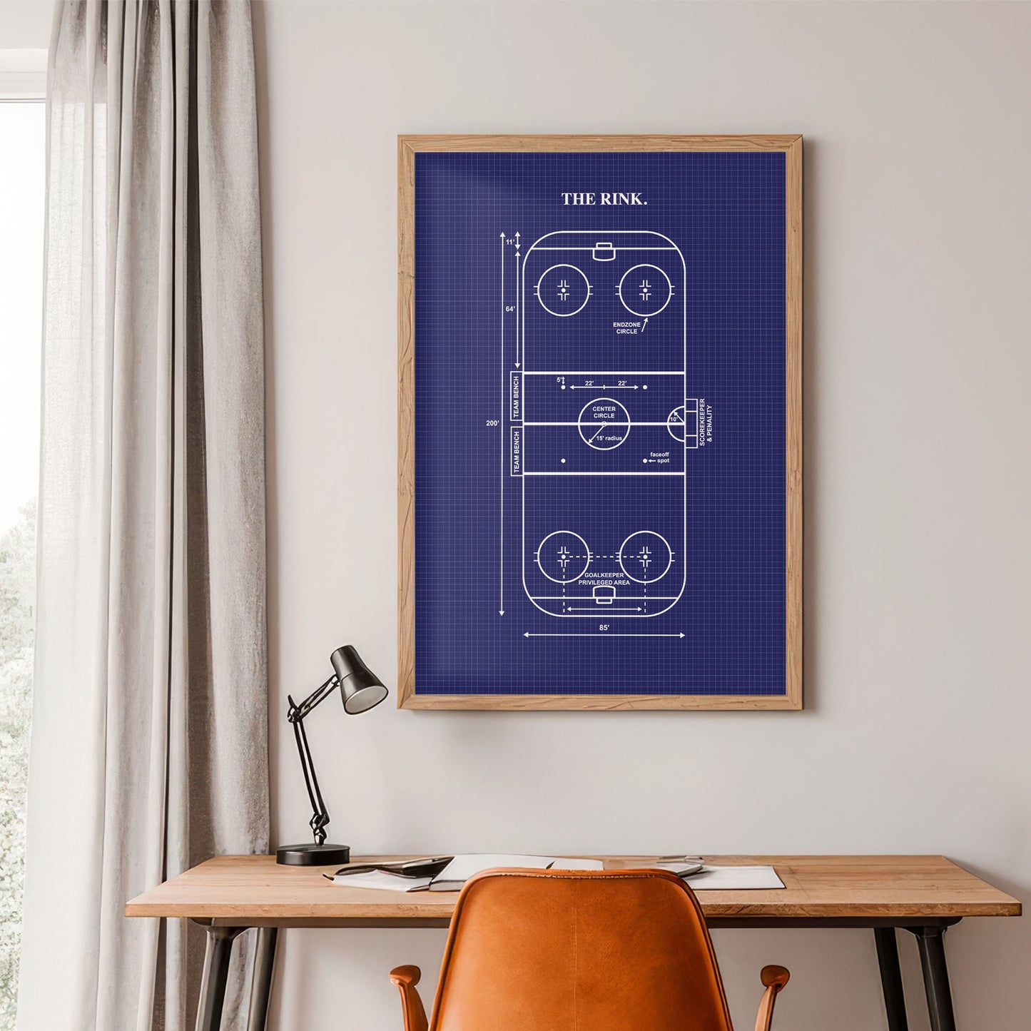 Framed blueprint of a rink on a wall above a desk with a lamp and chair.