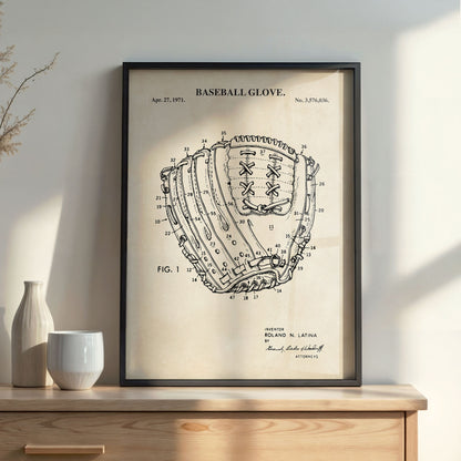 A framed vintage patent-style baseball glove print leaning on a wooden cabinet against a light wall, with a ceramic vase and small bowl beside it.