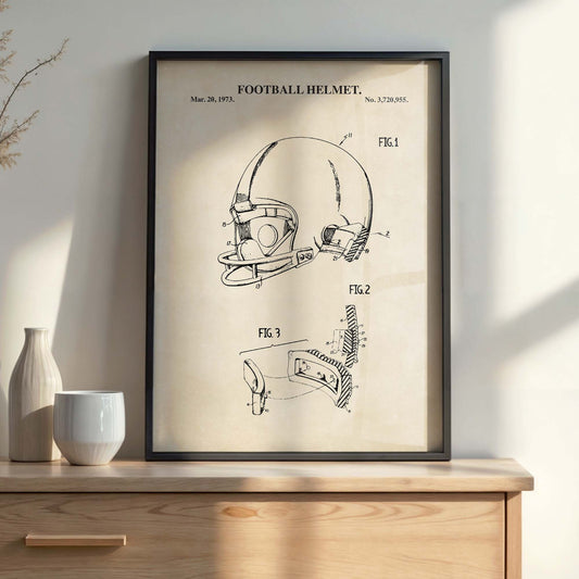 A framed vintage-style patent print of a football helmet on a light wood console table, with a ceramic vase and white cup beside it against a beige wall.