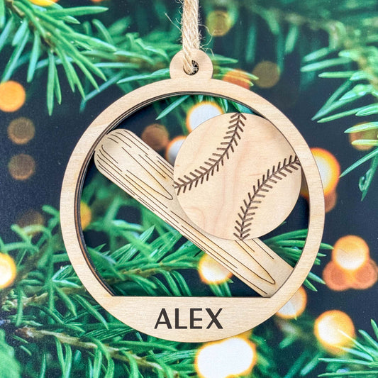 A personalized wooden baseball ornament hanging from a string, featuring an engraved baseball and bat design inside a circular frame with the name 'ALEX' at the bottom, set against a festive pine tree background with warm bokeh lights.