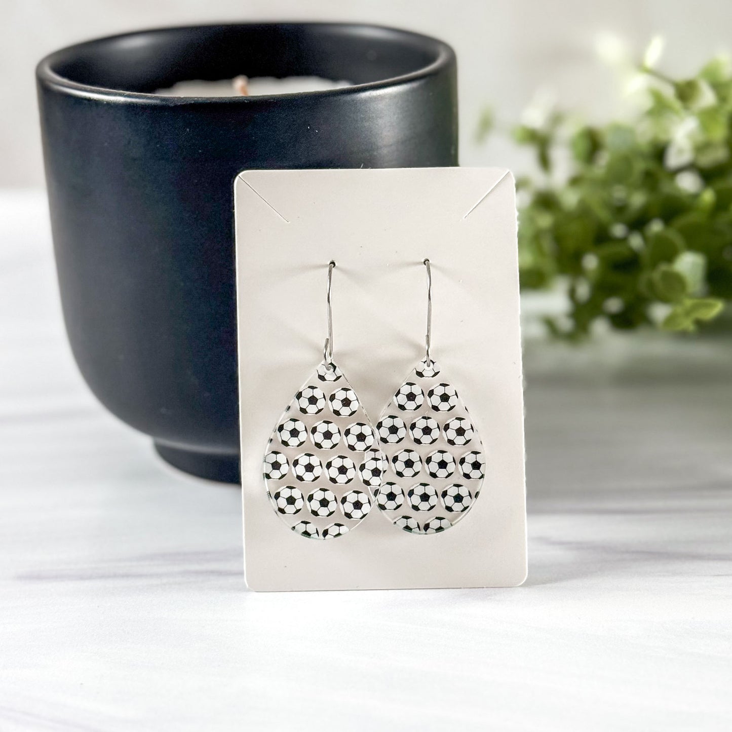 Pair of soccer ball earrings on a white card, with a dark candle and greenery in the background.