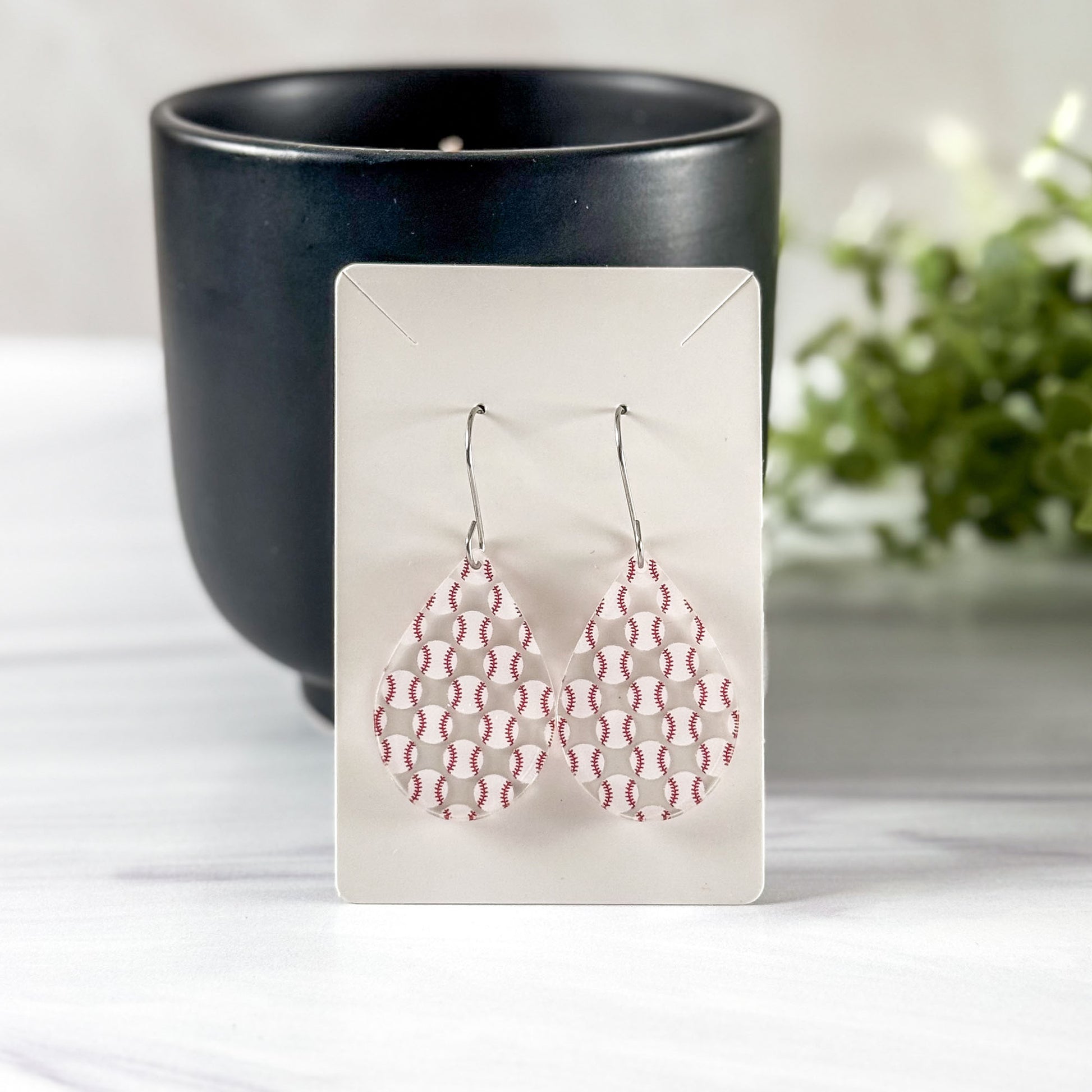Pair of red and white baseball earrings on a display card with a black cup and plant in the background.