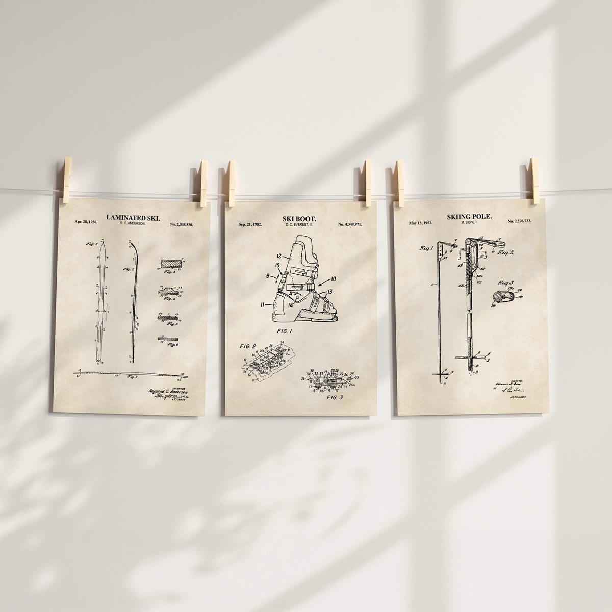 Three vintage-style patent art prints for snow sports are hung on a white wall by wooden clothespins. The prints feature black technical drawings on aged beige paper, including a laminated ski, ski boot, and ski pole.