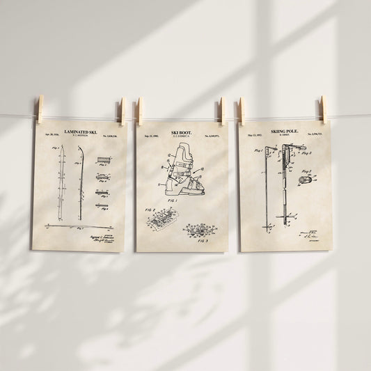 Three vintage-style patent art prints for snow sports are hung on a white wall by wooden clothespins. The prints feature black technical drawings on aged beige paper, including a laminated ski, ski boot, and ski pole.
