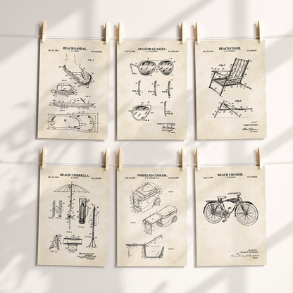 Six vintage-style beach-themed patent art prints are clipped to a white string against a sunlit wall. The prints feature black technical drawings and text for a beach sandal, aviator glasses, beach chair, beach umbrella, wheeled cooler, and beach cruiser, all on warm cream paper.
