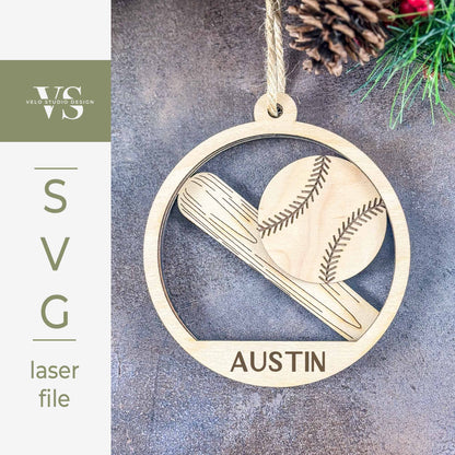 A laser-cut wooden baseball ornament design shown on a textured gray background with pine needles and a pinecone. The circular ornament features an engraved baseball and bat with the name 'AUSTIN' at the bottom.