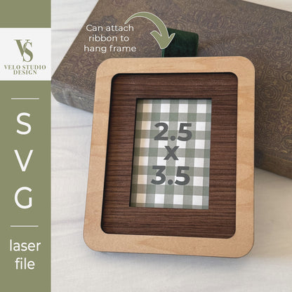 A laser-cut wooden picture frame with a light natural wood border and a darker wood inner section, shown on a tabletop with a decorative book in the background. A green ribbon is attached to the top.