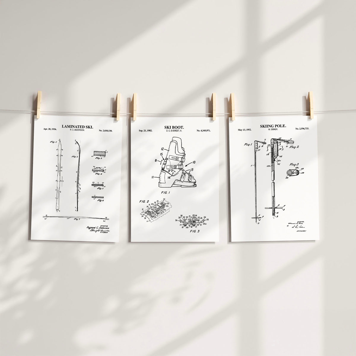 Three white patent-style art prints clipped to a string on a light wall, showing technical drawings labeled for skiing gear.