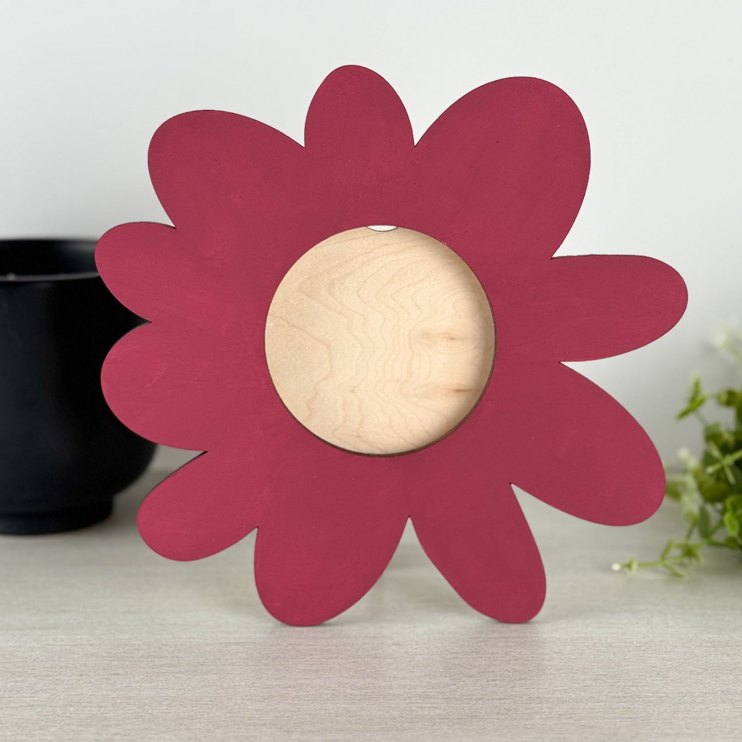 A flower-shaped wooden decorative piece with a dark red-pink painted petal border and a natural light wood grain center, standing on a light gray tabletop.