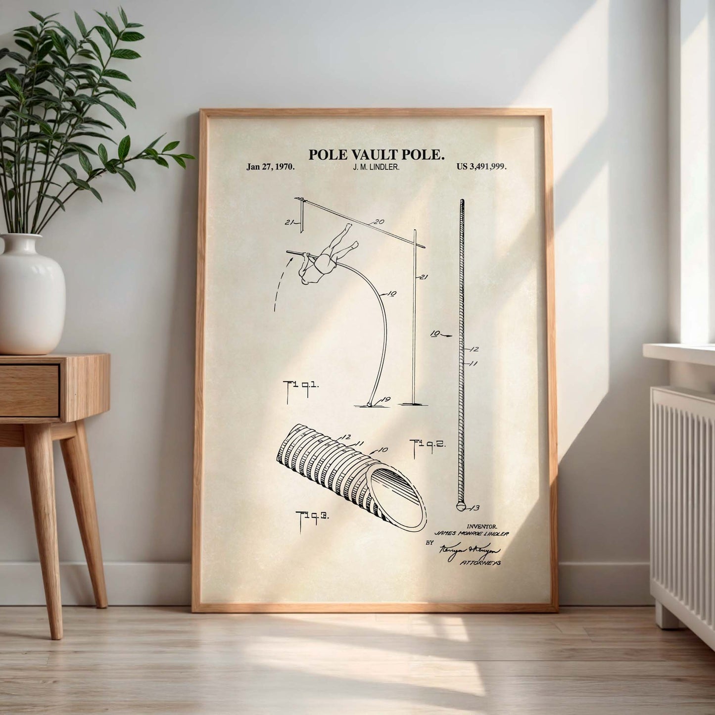 Framed patent poster of a pole vault pole on a wall in a room with a plant and wooden side table.