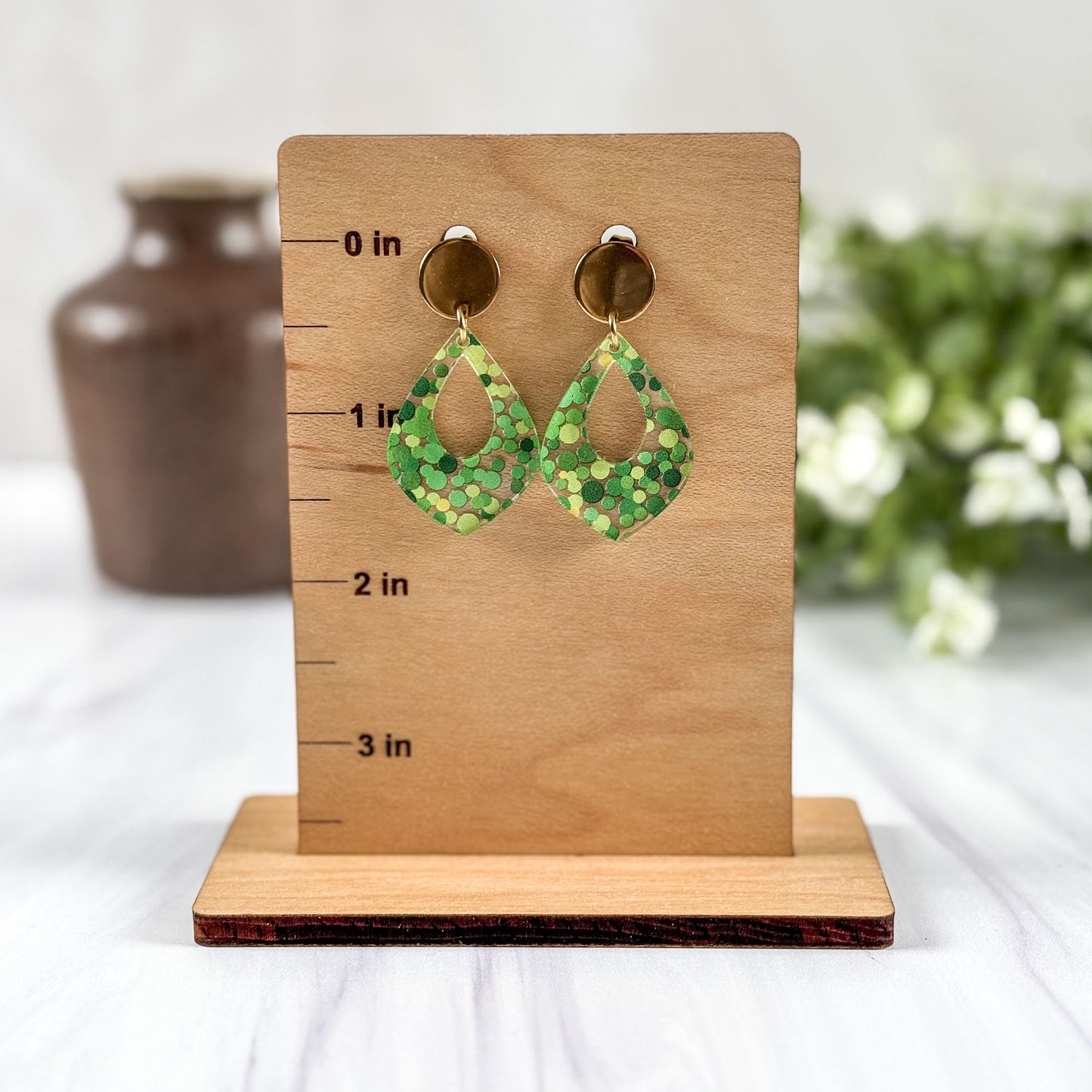 Green patterned earrings on a wooden stand with measurements, against a neutral background.