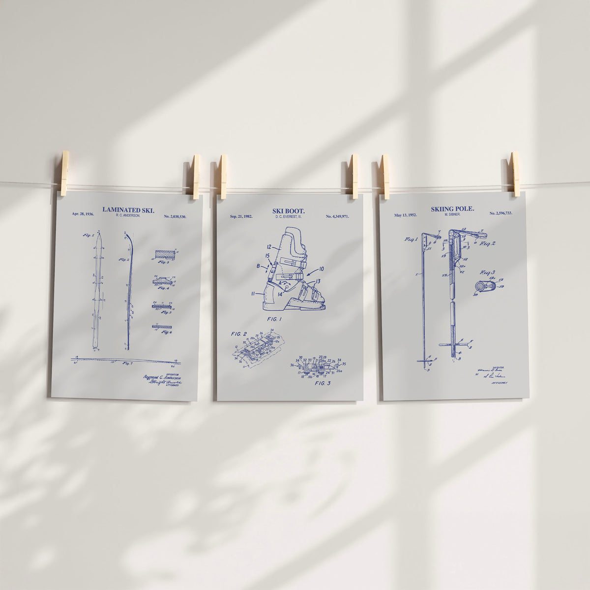 Three white patent-style art prints with blue technical skiing drawings are clipped to a string against a white wall, with soft shadow patterns cast across the scene.