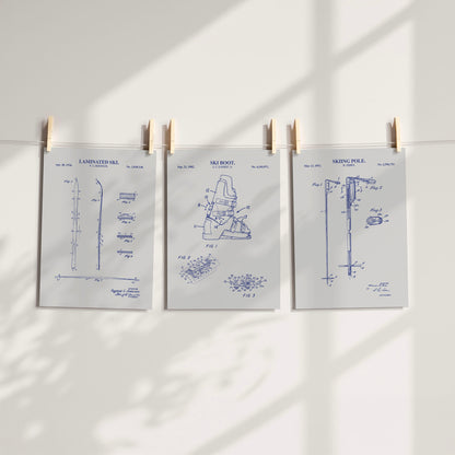 Three white patent-style art prints with blue technical skiing drawings are clipped to a string against a white wall, with soft shadow patterns cast across the scene.