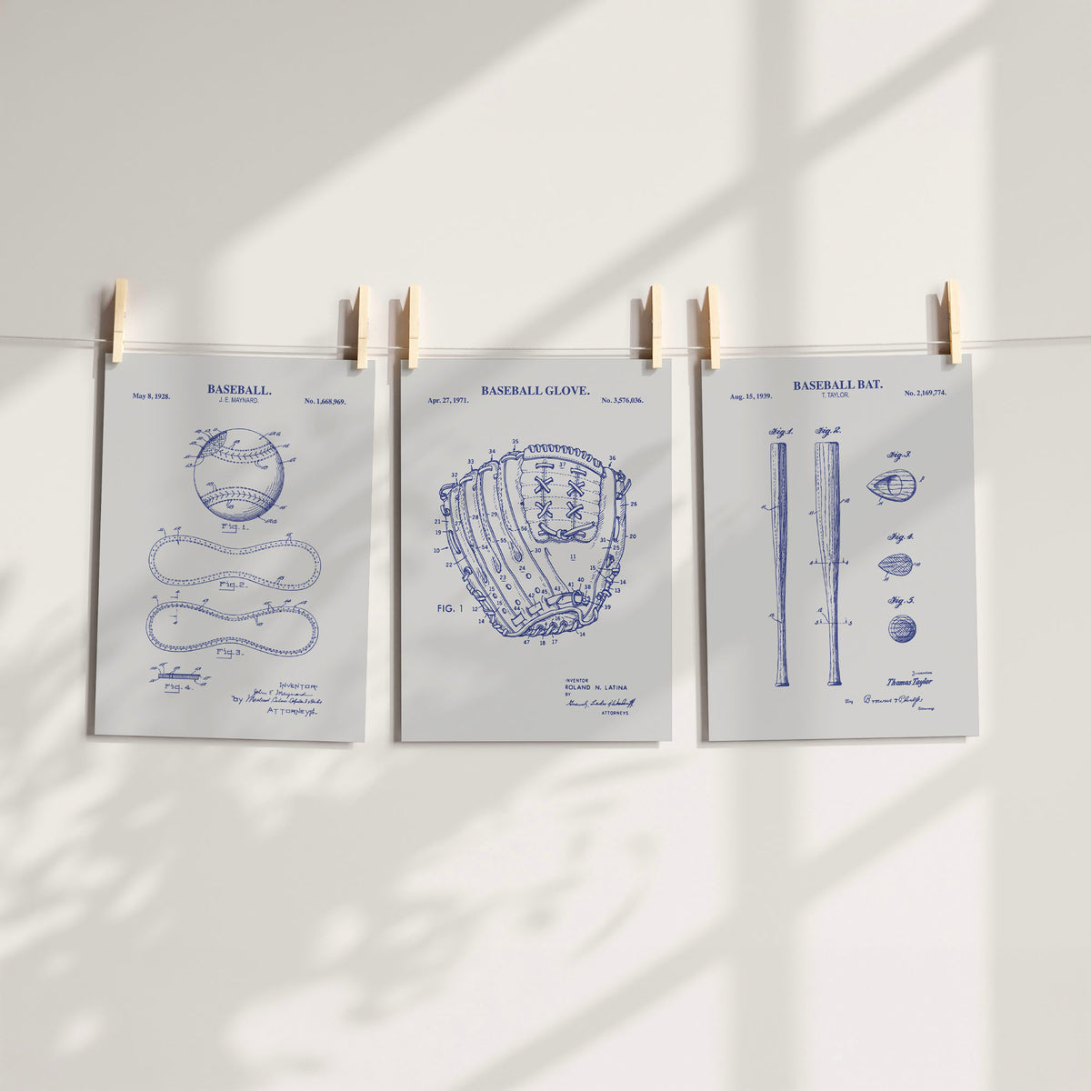 Three gray and navy baseball-themed patent art prints clipped to a string against a white wall with soft window shadows.