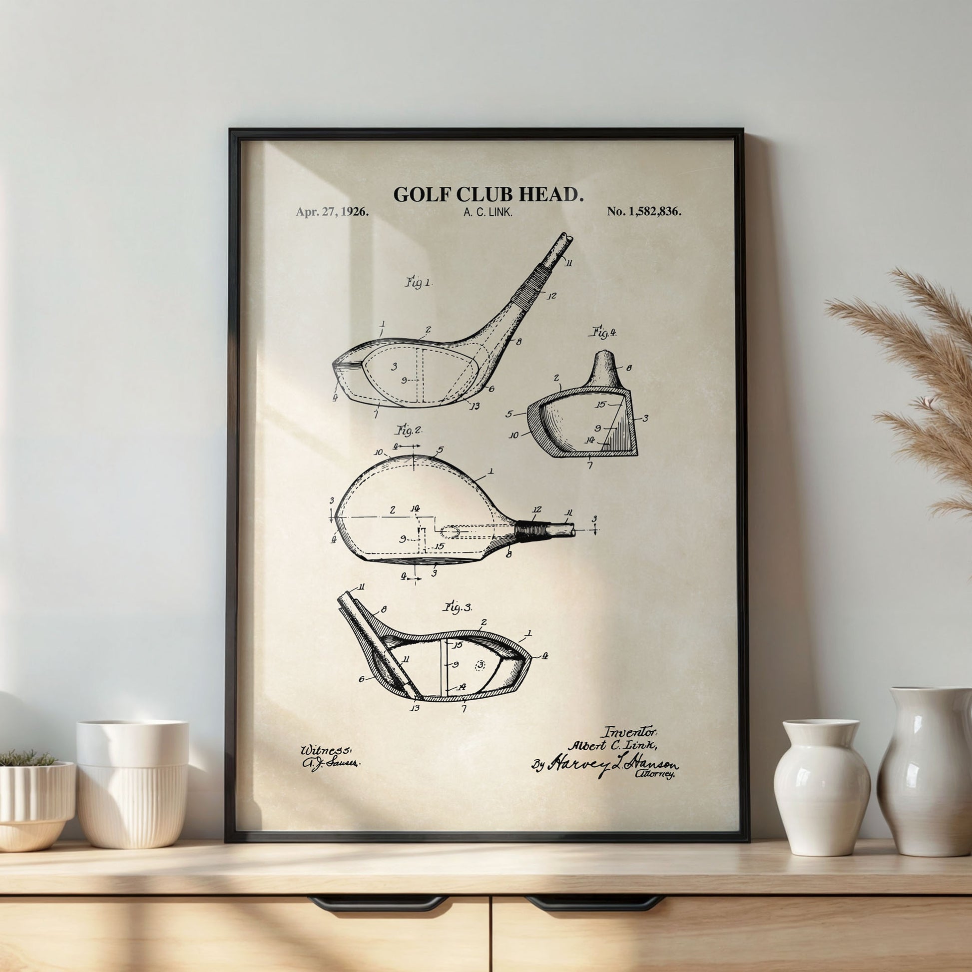 Framed golf club head patent print on a wall above a wooden cabinet with decorative vases.