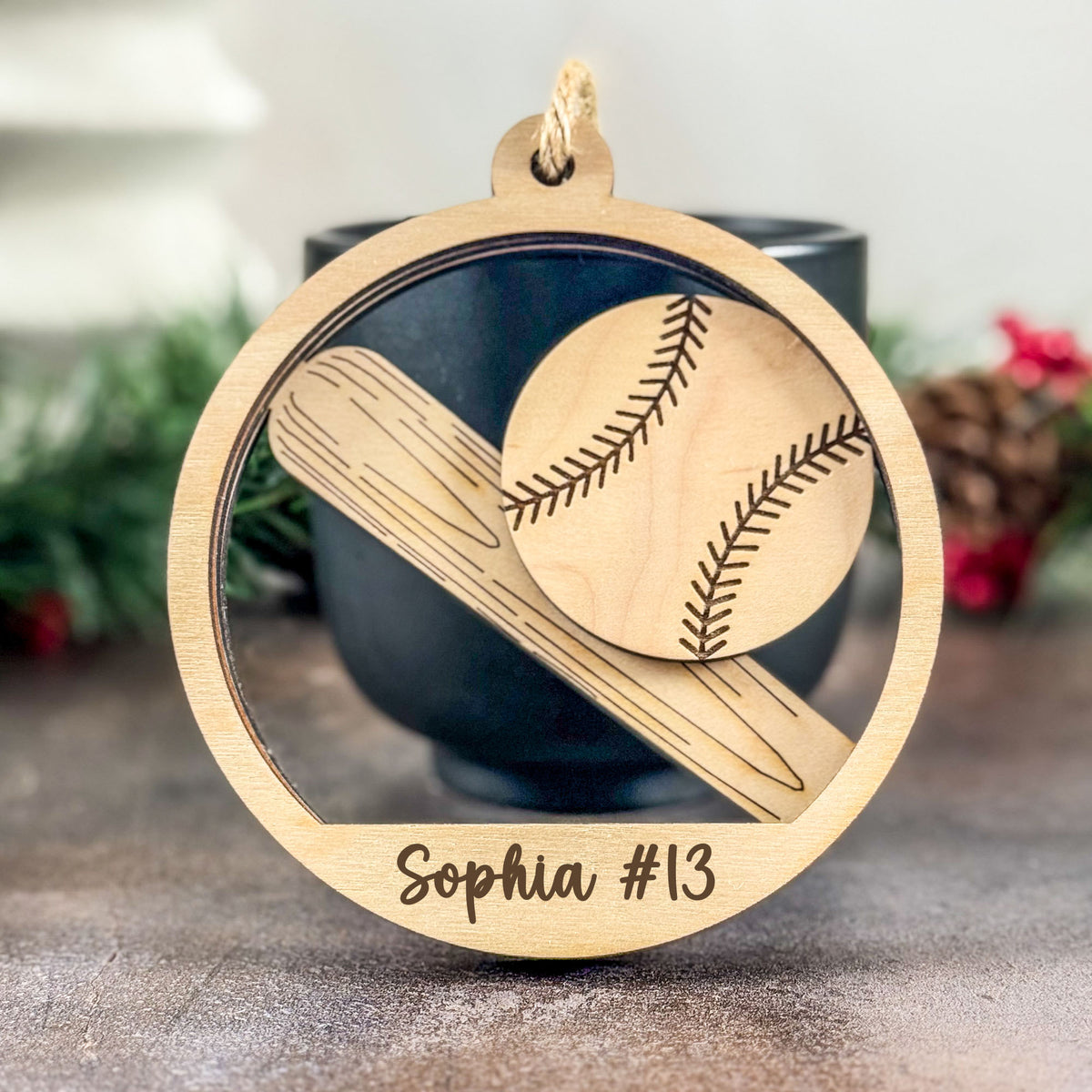 A personalized wooden baseball ornament hanging in front of a dark cup. The circular light wood ornament features an engraved baseball and bat design with the name 'Sophia #13' written at the bottom.