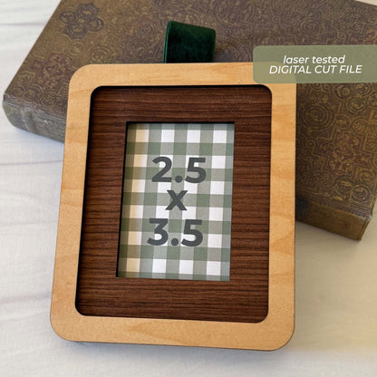 A square wooden picture frame with a light natural wood border and a dark wood inner mat, shown on a white surface with an old book in the background. A green label in the corner reads 'laser tested DIGITAL CUT FILE'.