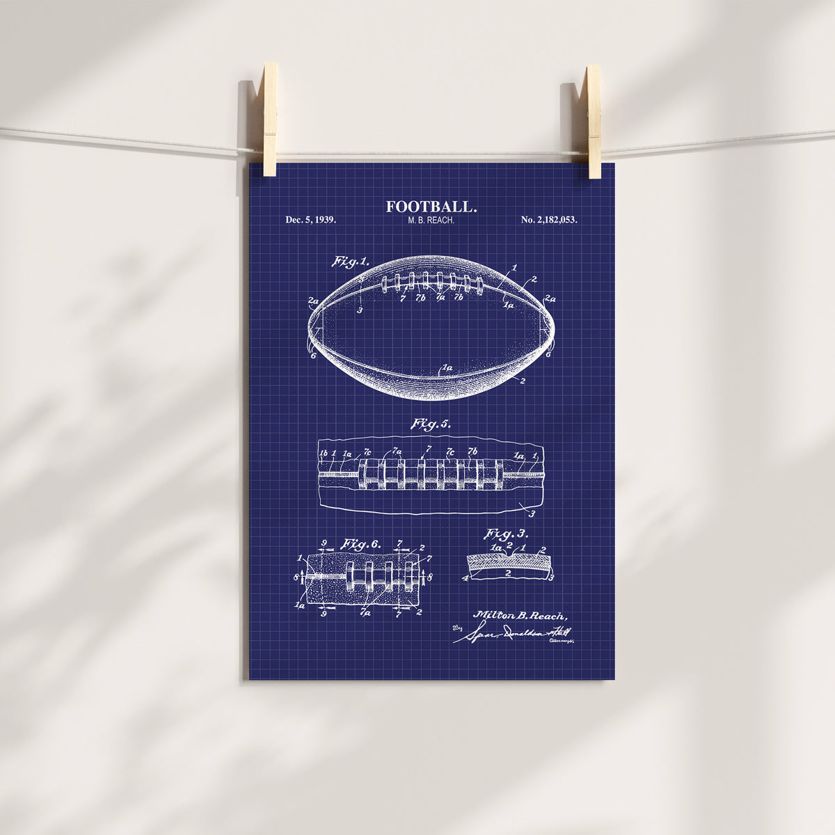 A navy blue poster featuring a vintage 1959 football patent drawing in white, hanging from two wooden clothespins on a white string against a white wall with soft leaf shadows.