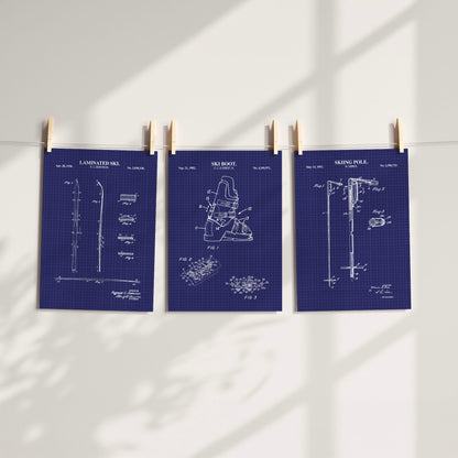 Three navy blue patent-style art prints of ski gear clipped to a string against a white wall, with soft window shadows cast across the scene.