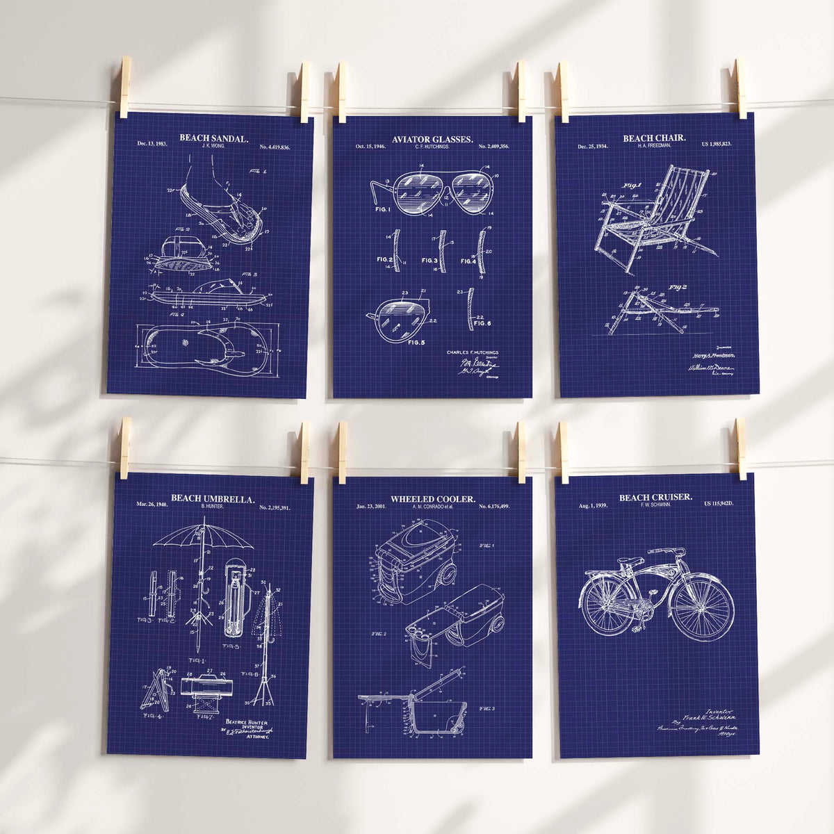 Six navy blueprint patent-style poster prints clipped to a string against a white wall, showing beach-themed inventions like a beach towel, hat, umbrella, cooler, and chair with white technical drawings.