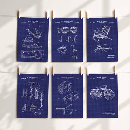 Six navy blueprint patent-style poster prints clipped to a string against a white wall, showing beach-themed inventions like a beach towel, hat, umbrella, cooler, and chair with white technical drawings.