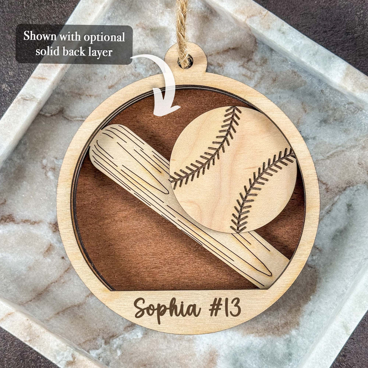 A personalized round wooden baseball ornament hanging on a marble tray. The ornament features laser-engraved designs of a baseball and bat, with the name 'Sophia #13' at the bottom.