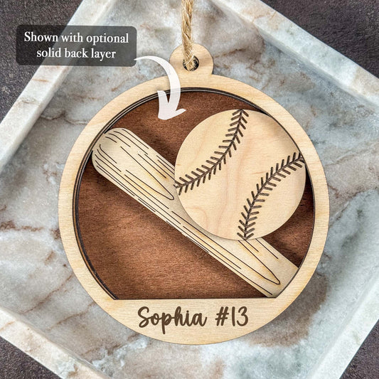 A personalized round wooden baseball ornament hanging on a marble tray. The ornament features laser-engraved designs of a baseball and bat, with the name 'Sophia #13' at the bottom.