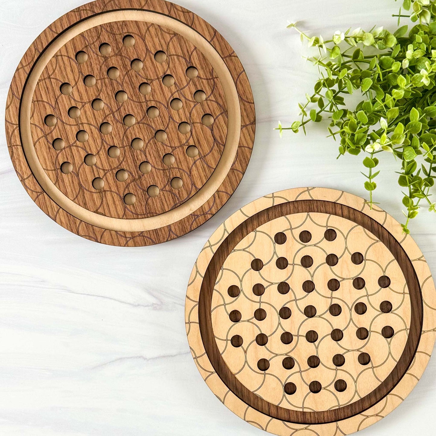 Two wooden marble solitaire games with a geometric pattern on a white surface with greenery.
