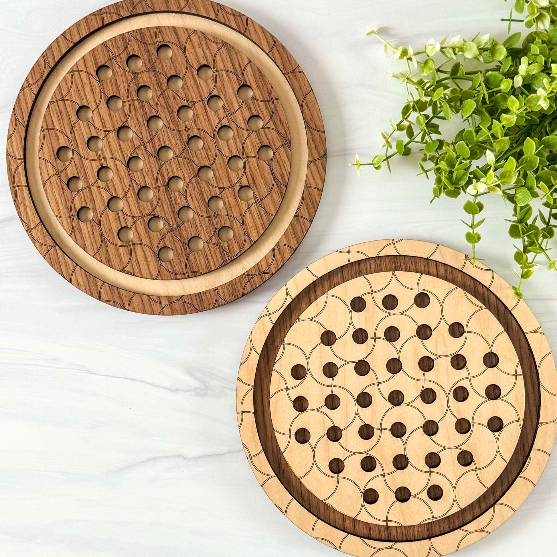 Two wooden marble solitaire games with a geometric pattern on a white surface with greenery.