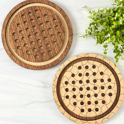 Two wooden marble solitaire games with a geometric pattern on a white surface with greenery.
