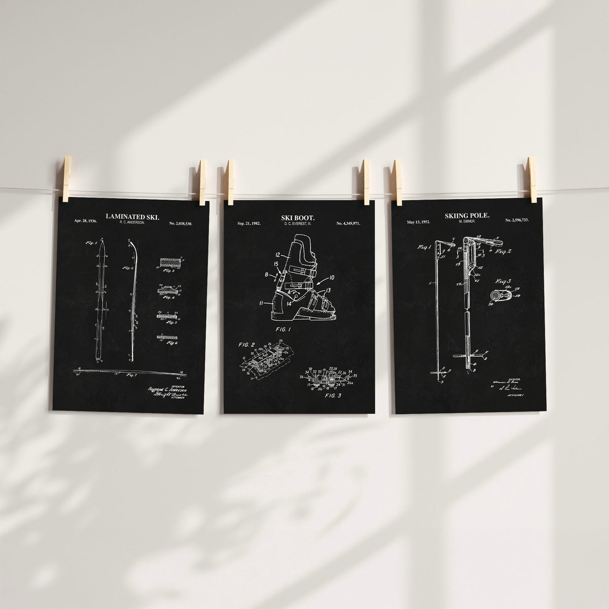 Three black patent-style art prints of ski gear with white technical drawings are clipped to a string against a white wall, with soft window shadows cast across the scene.