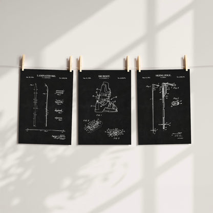 Three black patent-style art prints of ski gear with white technical drawings are clipped to a string against a white wall, with soft window shadows cast across the scene.