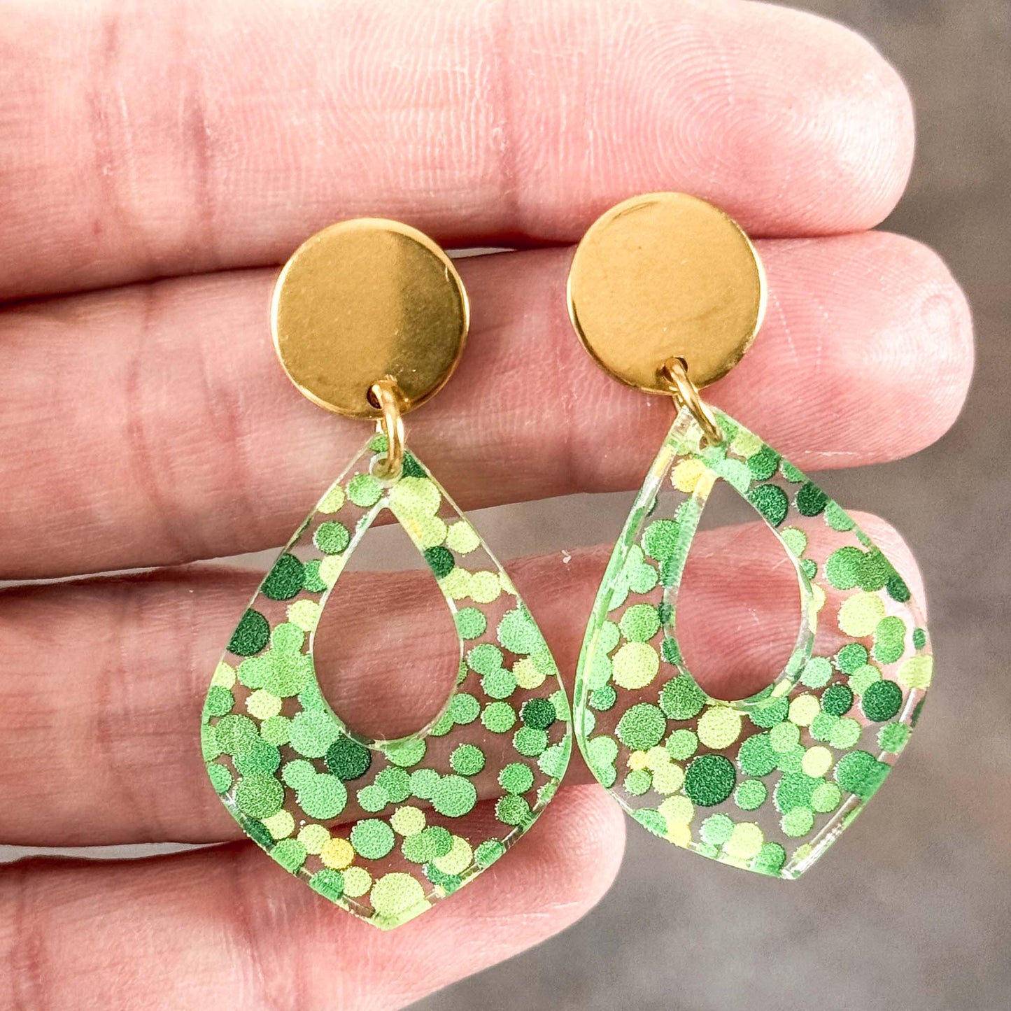 Gold earrings with green and yellow polka dot teardrop designs held in a hand.