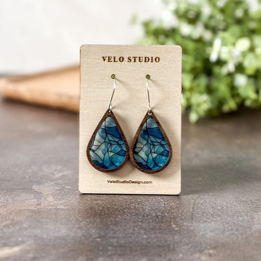 Teal and brown teardrop earrings on a Velo Studio branded card with a blurred green plant in the background.